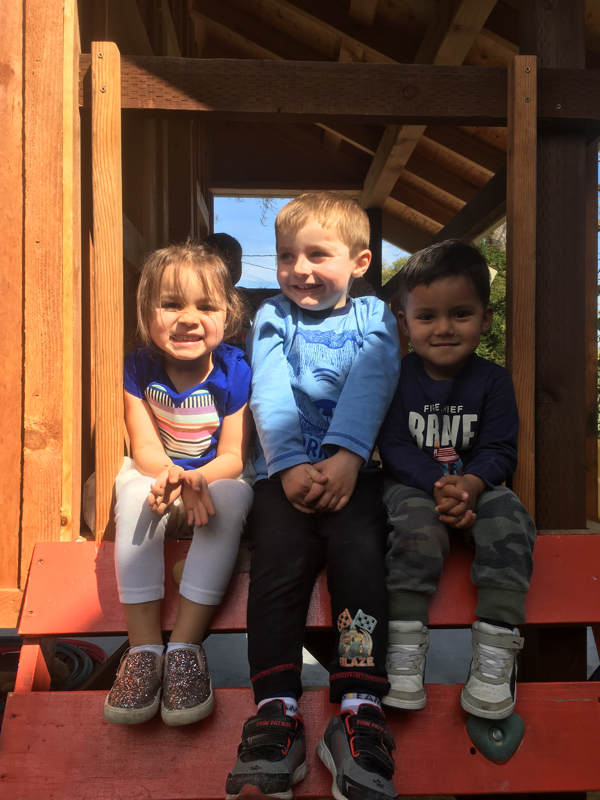 Three children smiling together on the wooden play structure