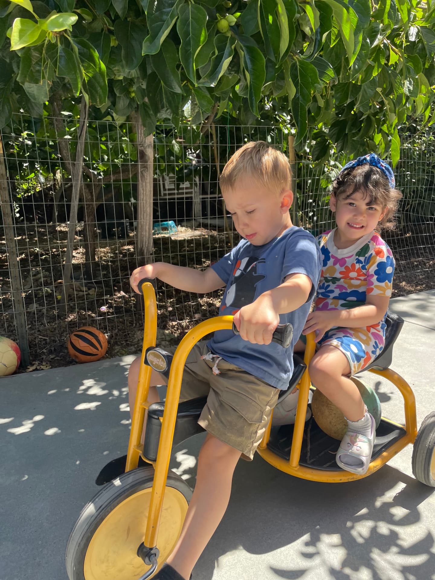 Two children sharing a ride on a yellow tricycle under the trees