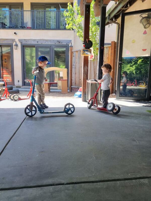 Two children riding scooters side by side in the outdoor play area